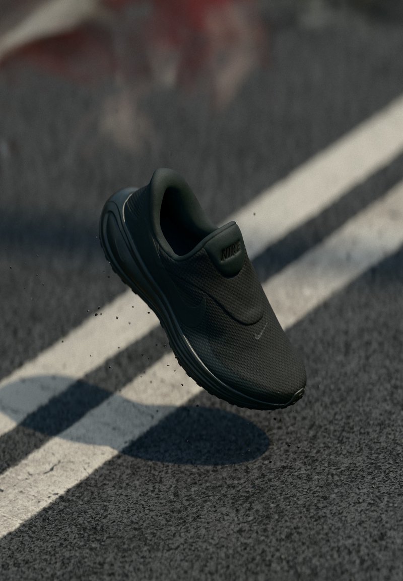 Μαύρο sneaker Nike slip-on που αιωρείται πάνω από ασφάλτινο δρόμο με λευκές διαγραμμίσεις, μικρά υπολείμματα στη σόλα ορατά.