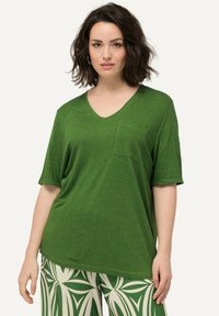Camiseta verde de cuello en V en una tela texturizada con un bolsillo en el pecho; corte holgado; combinada con pantalones estampados con diseños de hojas en verde y blanco.