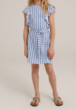 Mädchen trägt ein blau-weiß vertikal gestreiftes Kleid mit gerafften Ärmeln und einem Stoffgürtel, steht barfuß vor einem schlichten Hintergrund.