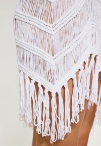 Witte franjes garment met een chevronpatroon, voorzien van textuur garen en lange kwastjes aan de onderrand, wat een gelaagd effect creëert.