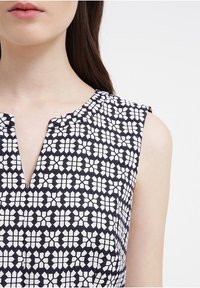 Laivastonsininen mekko, jossa on valkoinen geometrinen kukkakuviot, hihaton muotoilu, V-pääntie ja sileä kangaspinta. Lähikuva yläruumiin alueesta.