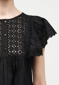 Μαύρο μπλουζάκι με κεντητό φλοράλ σχέδιο με διακοσμητικές οπές στο μέρος του κορμού, εμπρόσθιο κλείσιμο με κουμπιά και κοντά φουσκωτά μανίκια με λοξά δαντελένια άκρα.