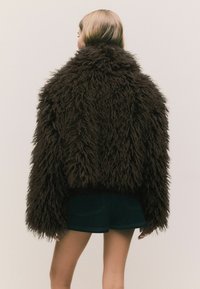 Person wearing oversized dark brown shaggy fur jacket and black shorts, shown from behind against a plain background.