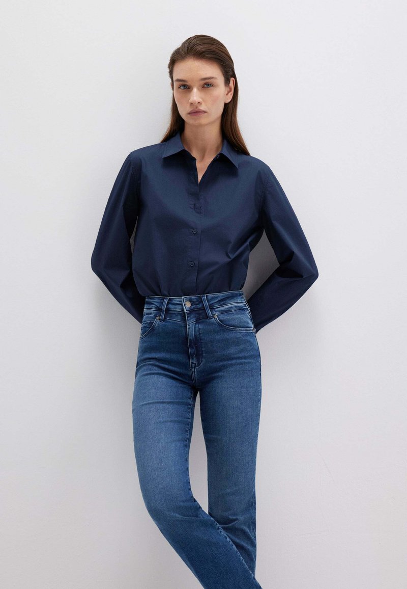 Camicia blu navy con bottoni e maniche lunghe, abbinata a jeans blu a vita alta e slim-fit; tessuto liscio con design classico e silhouette sartoriale.