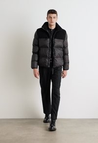 Giubbotto a soffietto nero con trapuntatura orizzontale e colletto alto. Presenta texture opache e lucide a contrasto, indossato sopra un completo nero.