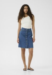 Jeans-A-Linien-Rock, mit Knopfverschluss, in mittellanger Höhe bis zum Oberschenkel, blaue Farbe, klassisches Design, getragen mit einem weißen Tanktop und schwarzen Flip-Flops.
