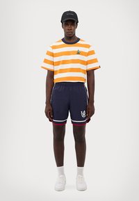 Orange og hvidstribet kortærmet skjorte, marineblå shorts med røde accenter, logo detalje, og hvide sneakers med ankelstrømper.