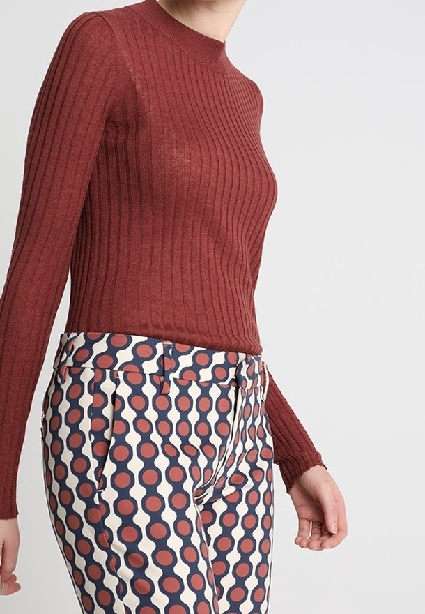 Femme portant un haut moulant à manches longues en tissu côtelé de couleur rouille et un pantalon taille haute avec un motif circulaire bleu marine, crème et rouille.