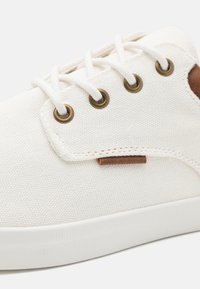 Canvas sneaker in white with brass eyelets, white laces, and a brown leather accent. Features a textured surface and rubber sole.