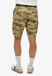 Mand med beige og grøn camouflage cargo shorts med knappede baglommer, hvide crew sokker og hvide sneakers med sorte accenter.