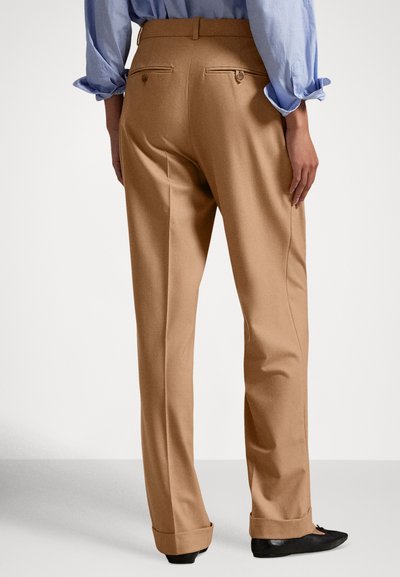 Παντελόνι ταγιέρ σε ταμπά χρώμα με λεία υφή, διαθέτει δύο πίσω τσέπες με κλείσιμο σε κουμπιά και ίσια γραμμή στο πόδι. Διακρίνονται μαύρα παπούτσια.