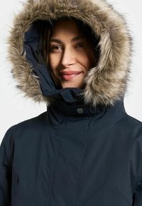 Tummansininen parka, jossa on turkisvuorattu huppu. Kangas on sileää, ja hupun reunoilla on pehmeä, pörröinen rakenne.