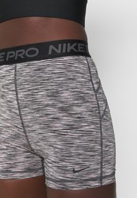 Spodenki Nike Pro w czarno-szarym wzorze w cętki, z szerokim czarnym pasem z napisem "NIKE PRO" oraz małym czarnym logo na brzegu.