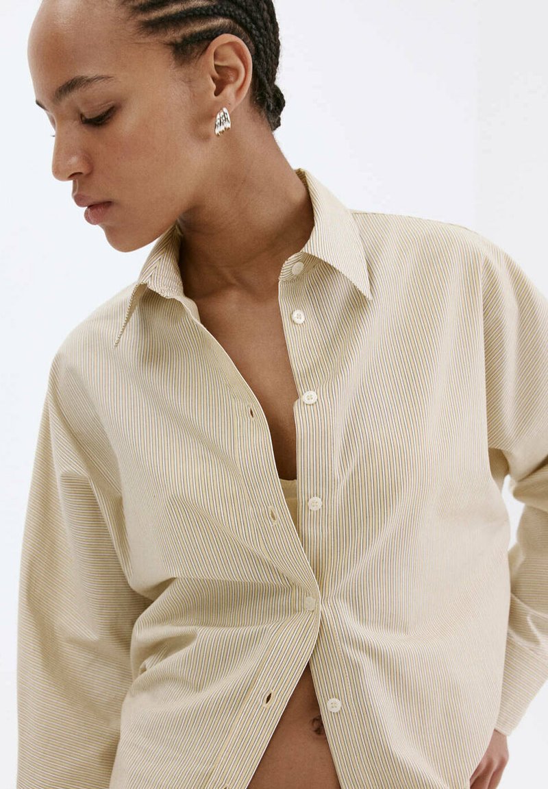 Femme aux cheveux tressés portant des boucles d'oreilles argentées et une chemise beige à fines rayures, regardant vers le bas, sur un fond blanc uni.