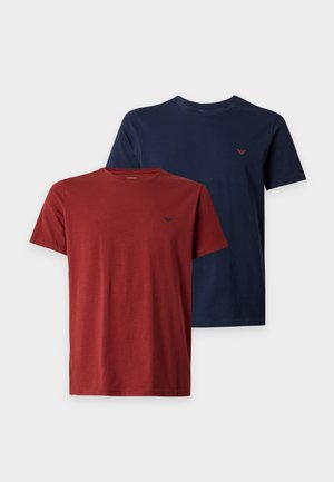 Junger Mann steht barfuß, trägt ein rotes kurzärmeliges T-Shirt und schwarze Hose vor einem schlichten weißen Hintergrund.