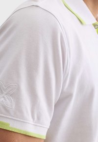 Biela polo košeľa s textúrovanou látkou, s jemne zeleným golierom a manžetami, a vyšitým logom na ramene.