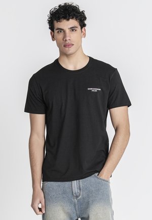 Joven con pelo rizado que lleva una camiseta negra "Gianni Kavanagh Atelier" y vaqueros azul claro, de pie con una mano en el bolsillo.