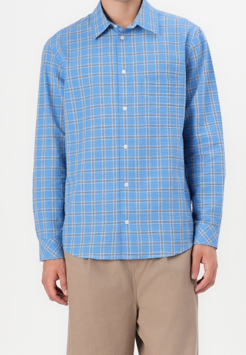 Homme portant une chemise à carreaux bleu clair à manches longues et un pantalon beige sur fond blanc.
