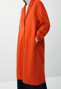 Cappotto in lana arancione con colletto a punte, vestibilità oversized e tasche laterali, caratterizzato da una texture liscia e lunghezza fino a metà coscia.