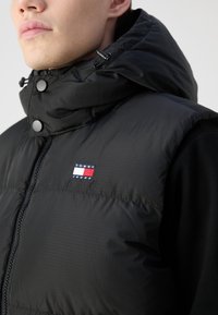 Crni puffer prsluk s visokim ovratnikom i kapuljačom. Ima zatvaranje na patentni zatvarač i sive dugmad za snap. Na prsima se nalazi mali višebojni logo.