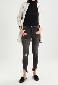 Ljusgrå stickad cardigan över en svart ribbad turtleneck, kombinerad med slitna grå skinny jeans och svarta plyschskor.