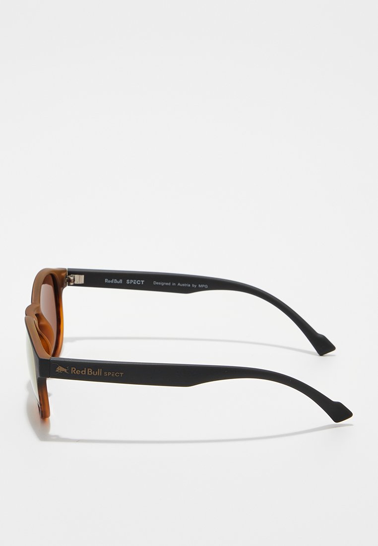 Okulary przeciwsłoneczne Red Bull SPECT z brązowymi przezroczystymi ramkami z przodu i czarnymi zausznikami, z logo i brandingiem Red Bull na zausznikach.