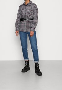 Persoon in een zwart-wit-rode geblokte jas met een zwarte heuptas, opgerolde blauwe jeans en zwarte gespen combatlaarzen, staande op een grijze vloer.