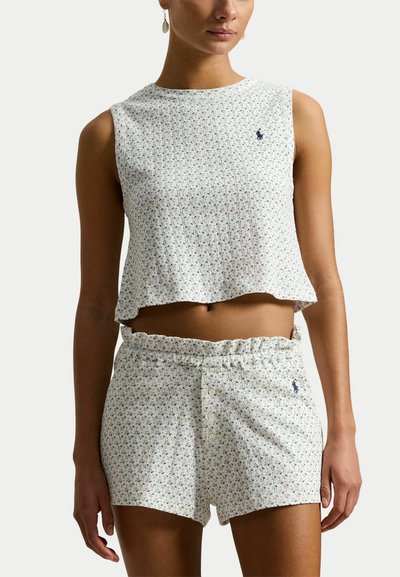 Femme portant un haut court blanc sans manches et un short taille haute assorti avec un petit imprimé floral et des logos brodés.
