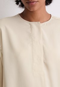 Beige, leichtes Hemd mit Rundhalsausschnitt und einer dezenten Knopfleiste. Glatte Textur mit feinen Nähdetails.