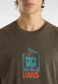 Olivově zelené bavlněné tričko s grafickým potiskem tváře panenky v modré a červené, s logem "VANS" tučně napsané červenou barvou pod ním.