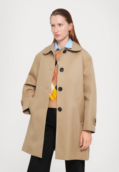 Cappotto oversize beige con colletto rotondo, bottoni neri e tasche laterali. Indossato sopra un maglione multicolore con un motivo a diamante.