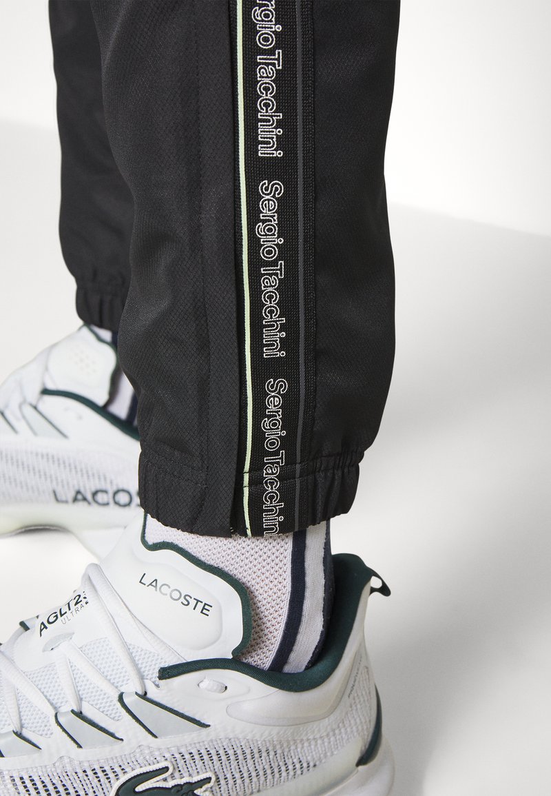 Pantalons de sport noirs avec le logo Sergio Tacchini sur le côté, associés à des chaussures blanches avec des accents verts et des chaussettes rayées.
