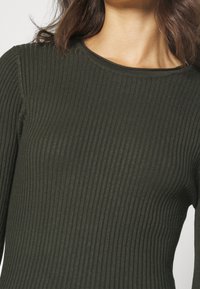 Mörkgrön ribbad långärmad topp med rund halsringning. Tyget har en mjuk textur och en figurnära passform som framhäver kroppens konturer.