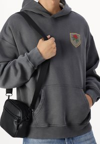 Szary hoodie z emblematem róży na piersi, w zestawieniu z małą czarną torbą na ramię, posiadającą zamki błyskawiczne i materiałowy pasek.