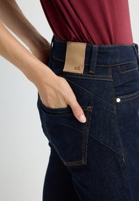 Džínové kalhoty v tmavě modré barvě s kontrastním prošíváním, s hnědou koženou nášivkou v pase a rukou vloženou v boční kapse.