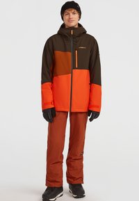 Oranssi ja ruskea vedenpitävä takki hupulla, jossa on vetoketjullinen rintatasku. Yhdistetty ruskeisiin housuihin ja mustiin hanskoihin, käytetään mustien saappaiden kanssa.
