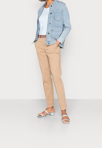 Kvinna som bär ljusblå denimjacka, blekblå topp, beige byxor med randig bälte och ljusblå klacksandaler mot vit bakgrund.