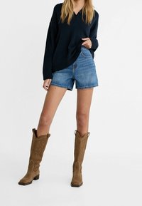 Marineblauwe oversized trui, blauwe denim shorts en tan suède laarzen tot aan de knie met decoratieve stiksels en een gestapelde hak.