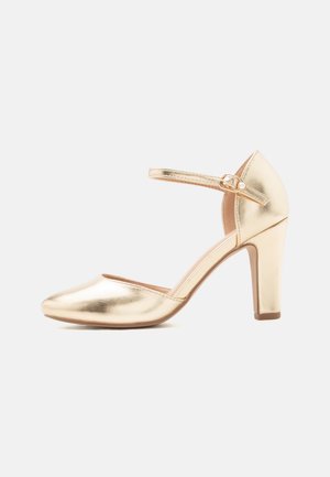 Zapato de tacón alto dorado con puntera redondeada, correa en el tobillo con hebilla pequeña y detalle de perla, y tacón ancho sobre fondo blanco.