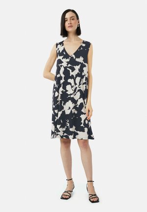 Robe marine à motif floral blanc, décolleté en V, sans manches, coupe ample et ourlet au genou. Portée avec des sandales noires à lanières.