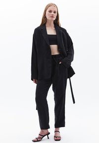 Černý lněný blazer s volným střihem a předním zapínáním na jeden knoflík, v kombinaci s krátkým topem a odpovídajícími kalhotami se šňůrkou na stažení. Černé sandály na vysokém podpatku.