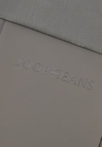 Tessuto grigio con una texture a coste è piegato su una superficie grigia uniforme, con un marchio in rilievo "JOOP! JEANS" per un aspetto elegante e moderno.