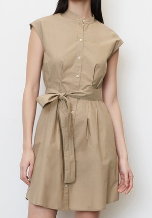Femme portant une robe-chemise beige sans manches avec une patte de boutonnage devant, un col mao et une ceinture à la taille, debout devant un fond uni.