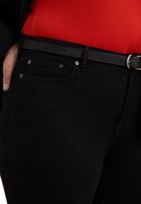 Sorte denim jeans med en glat tekstur, som har en forlommer og en metallknap. Sammen med et sort bælte og en rød skjorte.