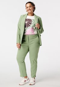Groen op maat gemaakt pak met een getailleerd colbert en bijpassende broek. De outfit heeft een wit shirt met bloemenpatroon en witte sneakers.