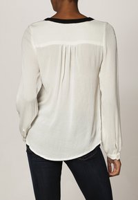 Blouse blanche à manches longues avec un col rond noir, tissu texturé, plis dans le dos et poignets à boutons, associée à un jean foncé.