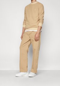 Beige texturovaná mikina s pružnými manžetami, přes bílý tričko. V kombinaci s béžovými kalhotami v uvolněném střihu a bílými teniskami.