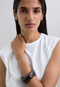 Bracelet rigide noir brillant sur un avant-bras bronzé, porté par-dessus un top blanc sans manches. Texture lisse et forme ronde, se fondant avec le ton de la peau.