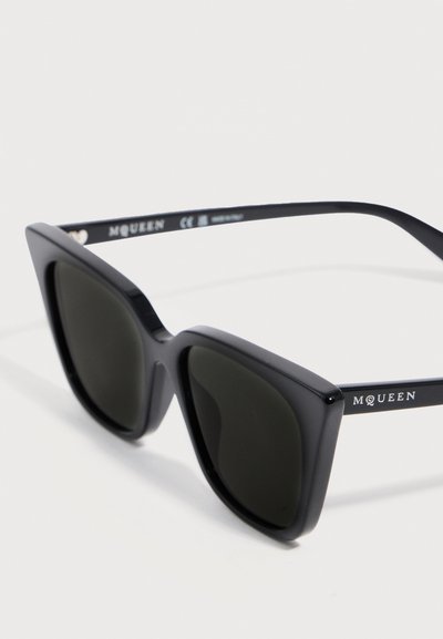 Μαύρα γυαλιά ηλίου με σχήμα γάτας και σκούρους φακούς, με το λογότυπο «McQueen» στο πλαϊνό μπράτσο, εμφανίζονται σε λευκό φόντο υπό γωνία.