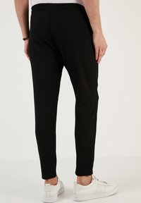 Čierne rebrované nohavice s elastickým pásom, slim fit, a detailom na zadnej vrecku, v kombinácii s bielymi teniskami na jednolatom pozadí.